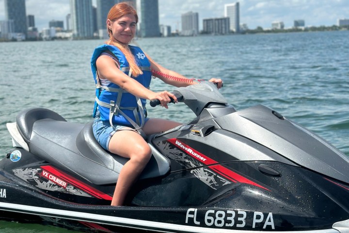 Person in blue life jacket riding a jet ski on a lake with cityscape in the background.